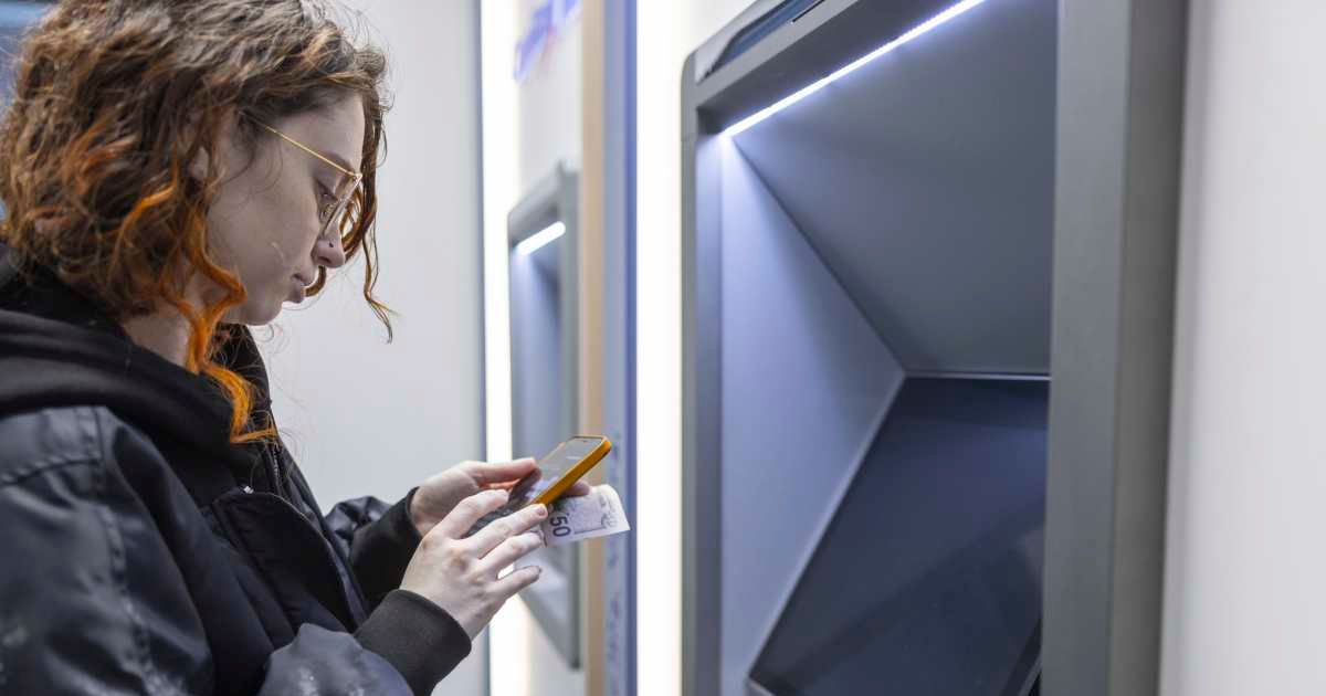Woman checking her phone for transaction updates while standing at an ATM (Cover image source: Getty Images | Alex Potemkin)
